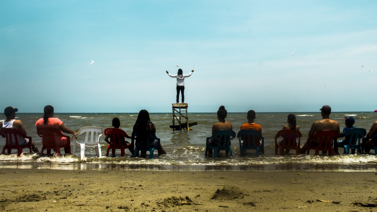 Un grupo de personas contempla desde la orilla de la playa a un hombre sobre una plataforma dentro del mar dirigiendo un concierto del mar